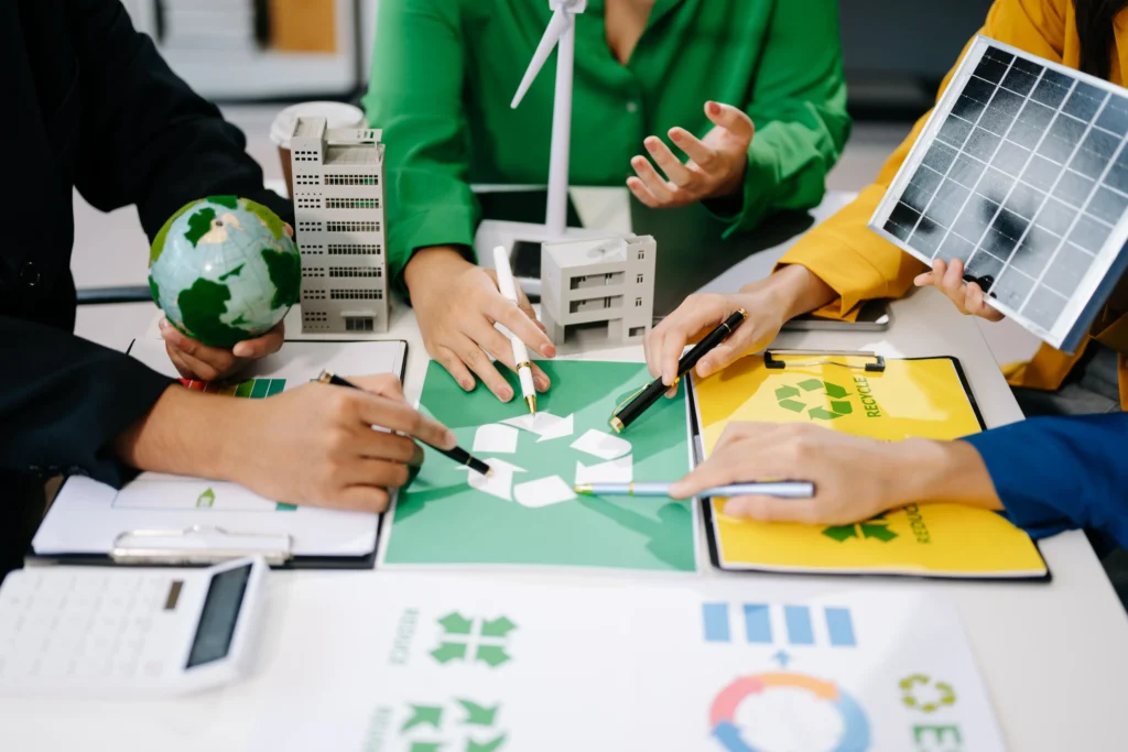 Personas diseñando estrategias de sustentabilidad con íconos verdes, edificios y recursos ecológicos en una reunión.